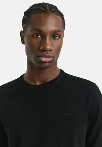 Pull en maille noir avec un col rond, présentant un logo discret sur la poitrine. Texture lisse, design ajusté, fabriqué en matériau léger.