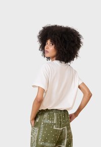 Cremefarbiges Baumwoll-T-Shirt mit kurzen Ärmeln, kombiniert mit olivgrünen gemusterten Hosen mit weißen Paisleymustern und Taschen.