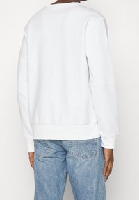 Osoba ubrana w prostą białą koszulę z długim rękawem i jasnoniebieskie dżinsy, stojąca tyłem do kamery na jednolitym tle.