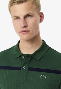 Grön polotröja med ribbad textur, tvåknappsknäppning och ett marinblått horisontellt rand. Har en liten Lacoste-logotyp på bröstet.