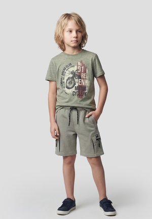 Niño rubio de pie con una mano en el bolsillo, vistiendo una camiseta gráfica de motocicleta verde oliva, pantalones cortos cargo a juego y zapatillas deportivas azul marino.