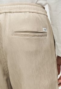 Pantalon en velours côtelé beige avec taille élastique et une poche fendue à l'arrière ornée d'une petite étiquette de marque rectangulaire blanche.