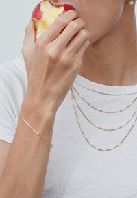 Gold and silver layered chains drape over a white shirt, accompanied by a delicate gold bracelet, held by a hand grasping a red apple.