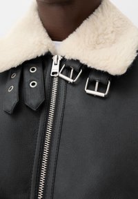 Veste en cuir noir avec un col en shearling crème, fermeture éclair argentée et détails en boucle, portée sur une chemise blanche.