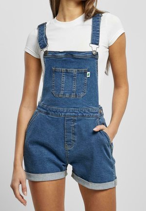 Mujer con peto corto de mezclilla azul con bolsillo frontal y camiseta blanca de manga corta, con una mano en el bolsillo, sobre un fondo liso.