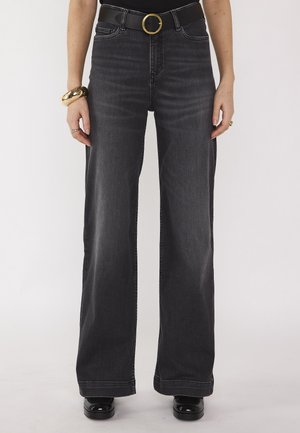 Persona che indossa jeans neri a vita alta e gamba larga con una cintura nera con fibbia dorata, scarpe nere e gioielli dorati alle mani.