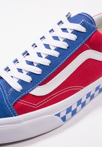 Vícebarevný plátěný teniska s modrými a červenými panely, bílými tkaničkami, bílým bočním pruhem a kostkovanou modro-bílou podrážkou.