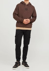 Brun huvtröja med en framficka och cirkulär logotyp, lager på lager med en beige skjorta. Buren med svarta cargobyxor och flertons sneakers.