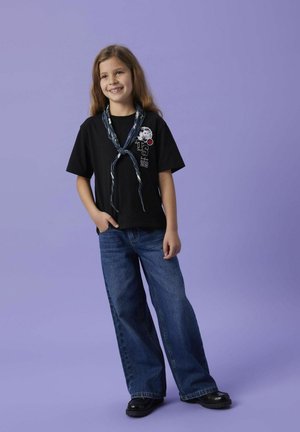 Svart kortärmad T-shirt med grafisk design och röd accent, i kombination med högmidjade blå jeans och svarta platåskor. Barnet står och ler.