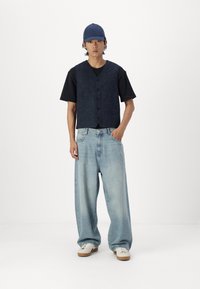 Mørkeblå denimvest med fem knapper, sammen med en sort t-skjorte og lyseblå brede jeans. Hvite joggesko fullfører antrekket.