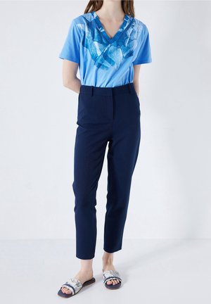 Kvinde iført en blå V-hals T-shirt med abstrakt print, navy smalle bukser og hvide mønstrede slidesandaler foran en ensfarvet baggrund.