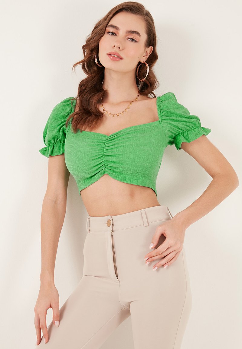 Top verde corto con tessuto testurizzato, scollo ampio e maniche a sbuffo; abbinato a pantaloni beige a vita alta con zip frontale.