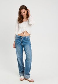 Biała koronkowa kurtka crop z wiązaniem z przodu, zestawiona z jasnoniebieskimi spodniami typu wide-leg. Obuwie ma różowe akcenty i białe sznurowadła.