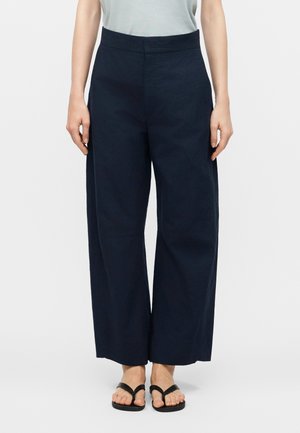 Γυναίκα που φοράει μπλε παντελόνι με ψηλή μέση και φαρδιά μπατζάκια, σε συνδυασμό με μαύρα σανδάλια δαχτυλίδι, στέκεται μπροστά σε ένα απλό λευκό φόντο.