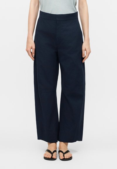 Femme portant un pantalon taille haute à jambes larges de couleur bleu marine, associé à des sandales noires avec anneau d'orteil, debout devant un fond blanc uni.
