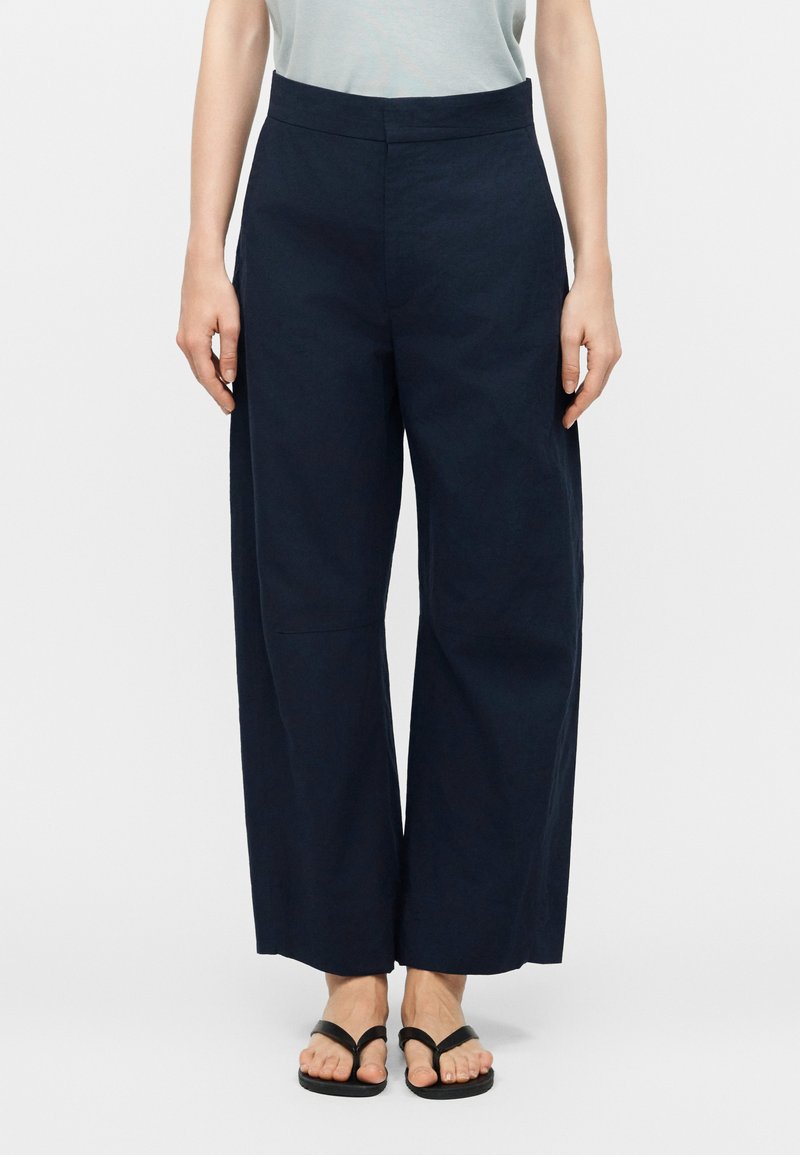 Γυναίκα που φοράει μπλε παντελόνι με ψηλή μέση και φαρδιά μπατζάκια, σε συνδυασμό με μαύρα σανδάλια δαχτυλίδι, στέκεται μπροστά σε ένα απλό λευκό φόντο.