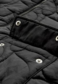 Détail d'une veste matelassée noire avec un tissu texturé, des boutons-pression en métal argenté et une fermeture éclair, ainsi que des motifs de couture matelassés.
