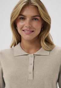 Béžová pletená polo košeľa s golierom, s gombíkovou légou a hladkou textúrou. Model má dlhé vlasy a neutrálny pozadí.