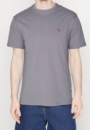 Mand iført en enkel grå t-shirt med korte ærmer med et lille rødt og hvidt flagemblem på venstre bryst og blå jeans.