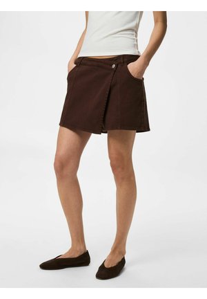 Femme portant un haut blanc sans manches, une jupe portefeuille marron avec des boutons devant, et des chaussures plates marron assorties, debout les mains dans les poches.