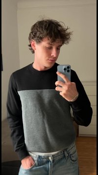 Giovane uomo con capelli ricci che indossa un felpa nera e grigia, mentre scatta un selfie allo specchio con uno smartphone in un ambiente interno semplice.
