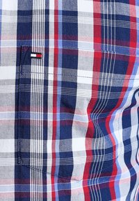 Nära bild av en rutig skjortficka med ett mönster i rött, vitt och blått samt en liten rektangulär flagg etikett.