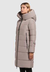 Béžový dlhý páperový kabát s veľkou kapucňou, horizontálnym prešívaním a zipsovými vreckami. Obsahuje kontrastný zips a logom detail na rukáve.