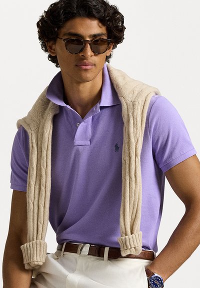 Μωβ πόλο μπλουζάκι με γιακά και λογότυπο. Μπεζ πουλόβερ με πλέξη καλωδίου ριγμένο στους ώμους. Ο καφέ ζώνη και το ρολόι χειρός είναι ορατά. Απαλό φόντο.