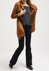 Braune Wildlederjacke über einem schwarz-weißen Punktenoberteil, kombiniert mit dunklen Schlagjeans und schwarzen Stiefeletten. Lässiges Outfit mit übereinander geschichteten Texturen.