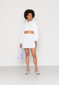 Top crop branco com detalhe em olho de perdiz e mangas bufantes, combinado com uma saia a condizer. Segurando uma mala lilás e usando sandálias com bloco de salto em lavanda.