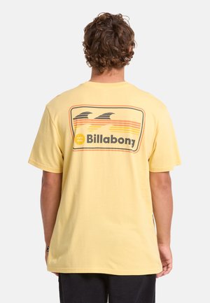 Uomo con capelli ricci che indossa una t-shirt gialla Billabong con una grafica onda retrò sulla schiena, in piedi davanti a uno sfondo semplice.