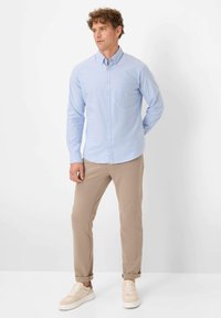 Hellblaues Hemd mit langen Ärmeln und einer Brusttasche, kombiniert mit beigen Hosen und hellen Sneakers. Flache Säume und schlichtes Design.