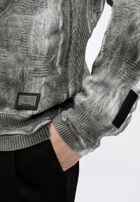 Gros plan d'une personne portant un pull gris texturé avec les mains dans les poches d'un pantalon noir. Le pull comporte des écussons de marque visibles sur la poitrine et la manche.