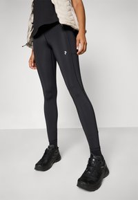 Czarne legginsy wykonane z elastycznego materiału, dopasowany krój, z detalem szwu bocznego i małym białym logo. Noszone z czarnymi sneakersami.