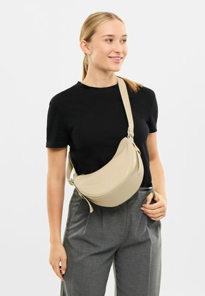 Beżowa skórzana torebka na ramię o zakrzywionym kształcie, z regulowanym paskiem i gładką fakturą, noszona na czarnej bluzce i szarych spodniach.