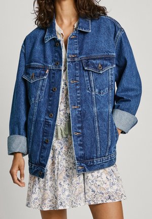 Femme portant une veste en jean bleue oversize sur une robe courte fleurie, avec les manches de la veste retroussées et une main dans la poche.