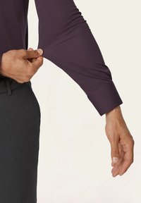 Dunkelviolettes Langarmshirt mit taillierter Passform, das dehnbare Stoffe zeigt. Eine Hand passt den Ärmel in der Nähe des Manschettenbereichs auf einem neutralen Hintergrund an.
