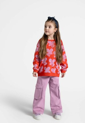 Czerwona bluza z wykrojem w różowe wzory motyli, połączona z lawendowymi szerokimi spodniami cargo z bocznymi kieszeniami. Białe sneakersy.