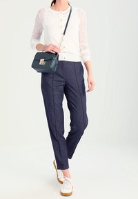 Vitt stickat kimono med mönstrade ärmar, marinblå byxor med vertikala sömmar och vita sneakers. Liten mörkgrön crossbody-väska.