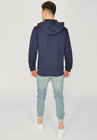 Marineblå hoodie med en baglomme og justerbar snor, parret med lysblå tapered jeans og hvide sneakers. Glat stofstruktur.