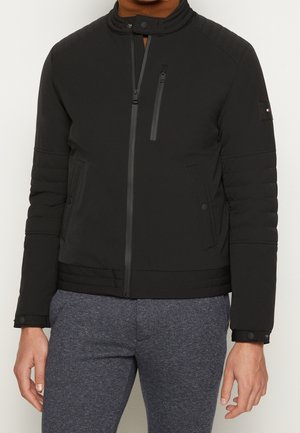 Veste isolante noire avec un col haut, des épaules matelassées, une poche poitrine zippée et des poignets boutonnés, associée à un pantalon de survêtement gris.