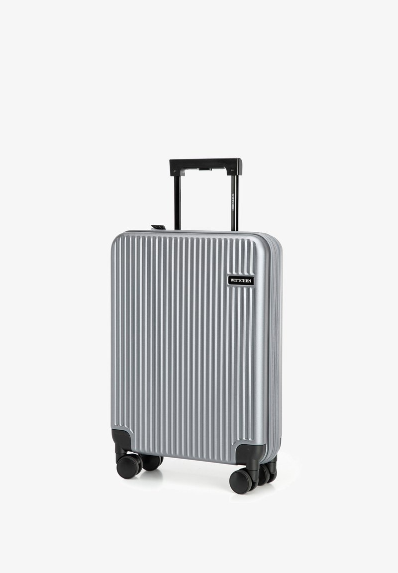 Valise rigide argentée avec des rainures verticales, accents de roues noires et une poignée rétractable. Étiquette de marque à l'avant.
