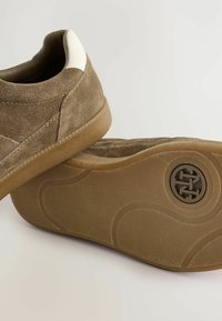 Braune Wildleder-Sneaker mit strukturiertem Obermaterial, kontrastierendem weißen Fersenakzent und einer Gummisohle mit einem kreisförmigen Logo und einem rutschfesten Muster.