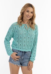 Maglione turchese lavorato a maglia con motivi in pizzo, maniche lunghe e neck tie a girocollo. Abbinato a pantaloni shorts in denim con bordi sfrangiati.