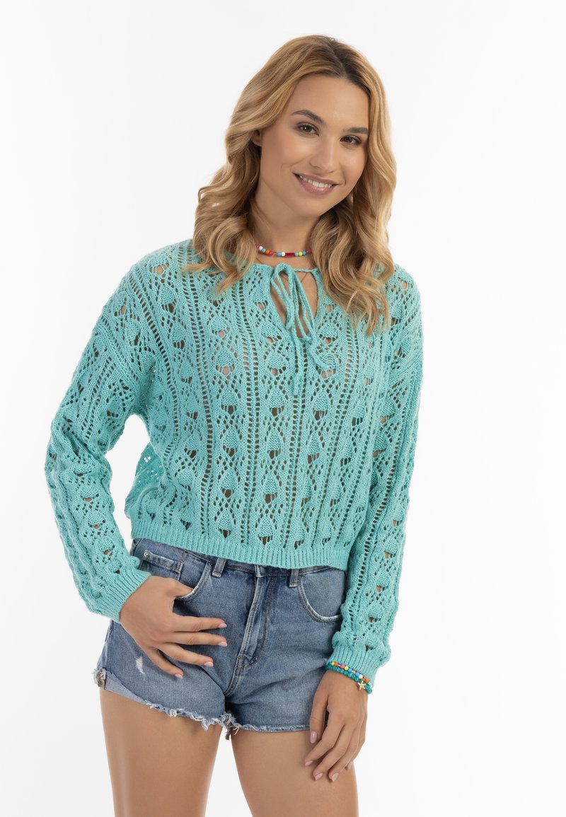 Maglione turchese lavorato a maglia con motivi in pizzo, maniche lunghe e neck tie a girocollo. Abbinato a pantaloni shorts in denim con bordi sfrangiati.