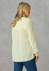 Blusa gialla chiara a maniche lunghe con dettaglio arricciato sul retro. Realizzata in tessuto morbido, presenta una vestibilità morbida e spacchi laterali per maggiore comodità.