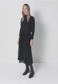 Musta pitkähihaisen yläosan yhdistelmä mustan, liehuvan hameen kanssa, jossa on läpikuultava kerros. Mustat nilkkurit viimeistelevät asun. Yksinkertainen, yksivärinen muotoilu.