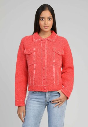 Jeune femme aux longs cheveux noirs portant une veste rouge vif boutonnée avec des coutures blanches et un jean bleu clair, debout devant un fond gris.