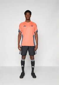 Športový outfit pozostávajúci z jasne oranžového dresu s krátkym rukávom a sivými akcentmi, doplnený čiernymi šortkami a čiernymi ponožkami s bielymi pruhmi.