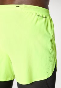 Primo piano di pantaloni sportivi giallo fluorescenti con leggings neri sotto, dotati di una piccola tasca con zip sul retro e fori di ventilazione.